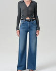 A model wearing a dark gray long-sleeve button-up shirt tucked into high-waisted, wide-leg blue jeans with a black belt, paired with black pointed-toe heels, against a light gray studio background.