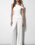 A woman modeling a white two-piece outfit consisting of an asymmetrical off-the-shoulder wrap top and matching wide-leg trousers, standing against a plain white wall.
