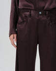 Close-up of a person wearing dark brown satin trousers with a matching top, showing the waist, belt, front pockets, and hand against a light gray background.