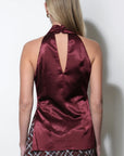 A woman shown from the back wearing a satin halter top in a deep burgundy color, paired with a patterned skirt, against a plain white wall.