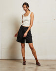 Woman wearing a white tank top and black shorts standing against a white wall front view