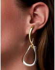 A close-up of a model's ear wearing a two-tone hoop earring with a polished gold top curve and a larger polished silver teardrop-shaped drop.
