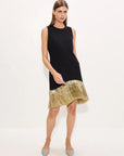 A woman modeling a sleeveless black dress with a flared gold hem, paired with pointed silver heels, against a plain white studio background.