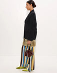 A model is shown in profile wearing a black blazer, wide-leg striped trousers in black, blue, yellow, and white, a textured gold skirt, and lime green pointed shoes, while holding a dark brown fan-shaped handbag against a plain white studio background.