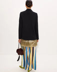 A model shown from the back wearing a black blazer, a gold sequined waistcoat, and a colorful vertically striped skirt in shades of blue, yellow, green, and brown. She is holding a dark brown handbag against a plain white studio background.