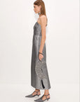 A woman shown from the side wearing a metallic silver spaghetti-strap midi dress with a textured pattern, holding a matching silver clutch, against a plain white studio background.