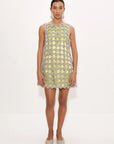 A model stands against a plain white studio background wearing a sleeveless, short dress covered in a metallic silver and pale yellow-green geometric mesh pattern with scalloped edges, paired with sparkly silver pointed shoes.