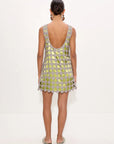 A woman viewed from the back wearing a sleeveless open-back mini dress covered in metallic gold and silver circular embellishments, set against a plain white studio background.