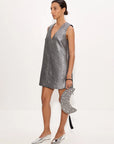 A woman models a sleeveless silver mini dress with a deep V-neck, paired with silver pointed-toe flats and a matching silver star-patterned crescent moon clutch bag, set against a plain white studio background.