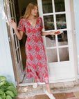 A woman wearing a red and white floral midi dress with short flutter sleeves and a V-neckline, standing in a doorway and holding a plate of tomatoes.