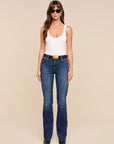 A woman modeling a pair of dark blue flared jeans with a black belt and gold buckle, paired with a white tank top and black sunglasses, against a solid beige background.