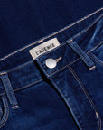 Close-up of dark blue denim jeans showing the waistband, button, belt loops, and a white fabric label that reads 'L'AGENCE'.