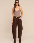 A woman modeling a pair of dark brown wide-leg cropped pants with a matching brown belt featuring a gold buckle, styled with a taupe camisole, sunglasses, and brown ankle boots against a solid beige background.