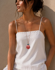 A woman wearing a white spaghetti-strap camisole top and matching bottoms, styled with a long red and blue \pendant necklace, leaning against a textured beige wall.