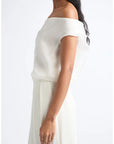 An off-the-shoulder white dress with a draped, layered neckline and a flowing skirt, shown on a model against a plain white background.