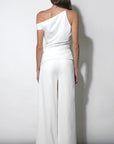 A woman shown from the back wearing a white one-shoulder wrap-style jumpsuit with wide-leg pants, standing against a plain light gray wall.