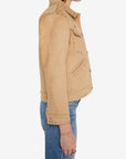 A woman shown in side profile wearing a cropped tan suede jacket with silver studs and blue jeans, photographed against a plain light gray studio background.