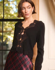 a model wearing a black cardigan with lace trim wth a plaid skirt standing in front of a window