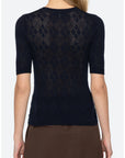 A woman shown from the back wearing a dark navy short-sleeve knit top with an openwork diamond pattern and brown bottoms, against a light gray studio background.