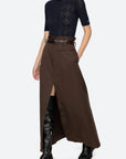 A model stands against a light gray studio background wearing a dark navy short-sleeve lace top, a long brown skirt with a front slit and belt, black knee-high boots, and black ankle boots.
