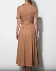A woman viewed from the back wearing a tan short-sleeve midi dress with a collared neckline and a tied waist, standing against a plain light gray wall.
