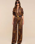 A woman modeling a leopard-print set with a high collar, long sleeves, and wide legs, styled with a black belt featuring a gold buckle, against a solid beige background.