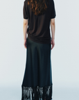 model wearing a black maxi skirt with fringe trim and a short sleeve brown tee on a white background, back view
