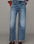 A model stands against a solid gray studio background wearing a cropped, long-sleeve striped knit polo shirt in rust red, navy, white, and light blue, paired with wide-leg blue jeans and dark brown leather loafers.