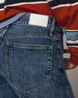 Close-up of the back waistband and pocket area of blue denim jeans, worn with a striped long-sleeve shirt. A blank white rectangular patch is visible at the back waistband.