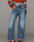 A model stands against a solid gray studio background wearing a cropped, long-sleeve striped knit polo shirt in rust red, navy, white, and light blue, paired with wide-leg blue jeans and dark brown leather loafers.