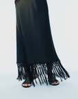 A black maxi skirt with a fringed hem, shown on a model wearing black strappy flat sandals against a plain white studio background.