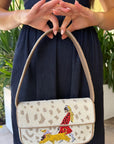 A person in a navy dress holds a New Staud Tommy beaded handbag with a leopard print pattern and a colorful embroidered figure on the front flap.