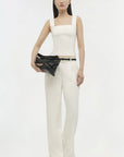 Woman wearing a white sleeveless top and pants with a black clutch against a white background
