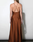 A woman viewed from the back wearing a long satin-like slip dress in a warm rust brown color, with thin crisscross back straps, standing against a plain light gray wall.