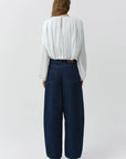 A woman viewed from the back wearing a white long-sleeve pleated blouse and dark blue wide-leg denim pants, standing against a light gray studio background.