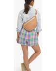 A woman shown from behind wearing a light blue denim shirt, pastel plaid shorts, and beige wedge sandals, with a woven orange and white crescent-shaped crossbody bag worn across her back.