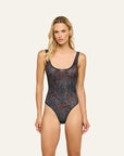 A blonde model wearing a bodysuit with a dark, abstract marbled pattern in black, blue, and brown tones, posed against a solid off-white background.