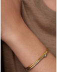 A close-up of a person's wrist wearing a slim two-tone metal bangle bracelet, featuring one polished gold-toned band and one silver-toned band, against a taupe knit top.