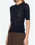 A woman wearing a dark navy blue short-sleeve knit top with a subtle argyle pattern, paired with brown trousers, against a light gray studio background.