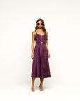 A woman modeling a sleeveless plum purple midi dress with a thin gold belt at the waist, standing against a seamless white studio background.