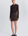 A model stands against a light gray studio background wearing a dark brown long-sleeve polo shirt, an olive green wrap-style mini skirt with a buckle detai, and black slip-on loafers.
