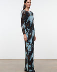A woman modeling a long-sleeve maxi dress with a blue, brown, and black abstract tie-dye pattern, standing in a minimalist studio with a white wall and light concrete floor.