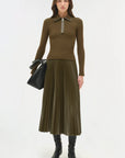 Woman wearing a brown/green sweater and pleated skirt with a black bag on a white background