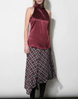 A model stands against a plain white studio wall wearing a sleeveless satin halter top in deep burgundy with a matching plaid midi skirt, black tights, and black heeled sandals.