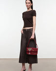A model stands in a studio wearing a dark brown short-sleeve crop top, dark brown wide-leg cropped pants, and black heeled sandals, while holding a dark red handbag with a cocktail glass detail.