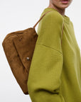 A woman wearing an oversized olive green knit sweater carries a brown suede shoulder bag on her back against a plain light gray studio background.