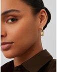 A close-up of a model wearing a gold chain-link drop earring, showing the earring against her ear. She is wearing a dark brown collared shirt against a light gray background.