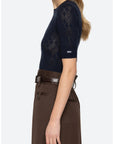 A woman shown from the side wearing a navy blue short-sleeve knit top with a textured argyle pattern and a small white logo on the sleeve, paired with a brown high-waisted skirt.