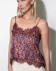 A woman modeling a burgundy floral lace camisole top with thin straps and a ruffled hem, paired with cream pants, against a plain white wall.