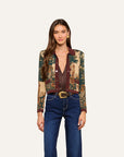 A model wearing a cropped, long-sleeve button-front cardigan with a detailed landscape tapestry print in beige, green, and brown, paired with dark blue jeans and a gold-buckled belt, against a solid off-white background.